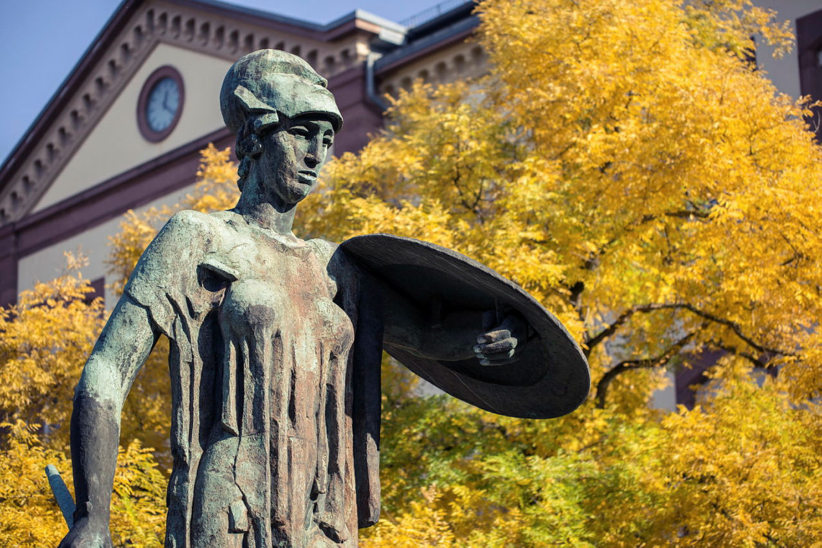 Bronzestatue einer Frau mit Helm und Schild vor einem Gebäude und gelben Herbstbäumen.