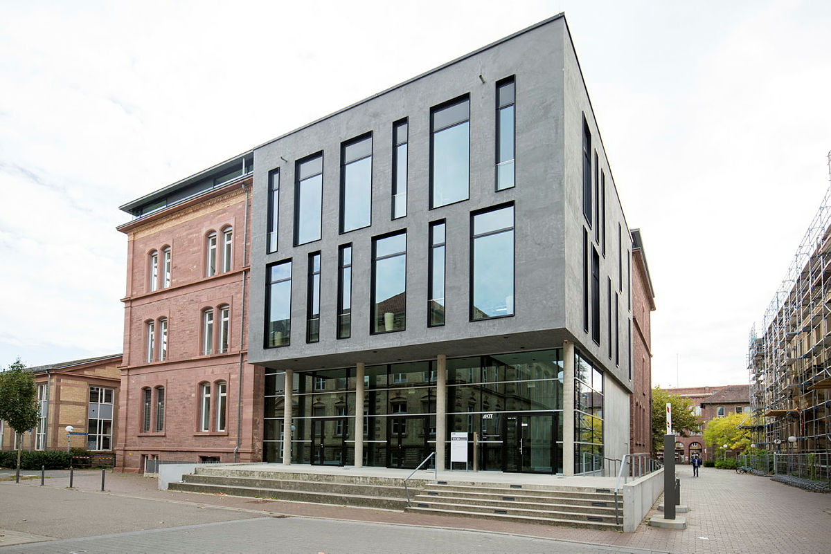 Moderner grauer Gebäudeteil neben einem historischen Sandsteingebäude mit großer Glasfront im Erdgeschoss.