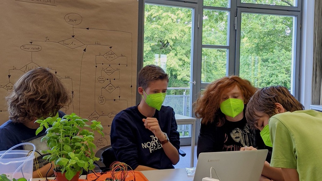 Schuelerinnen und Schueler mit Maske am Tisch mit Elektronik