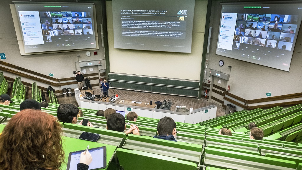 Sicht auf großen Hörsaal aus oberer Reihe mit gleichzeitiger Online-Konferenz auf drei großen Bildschirmen